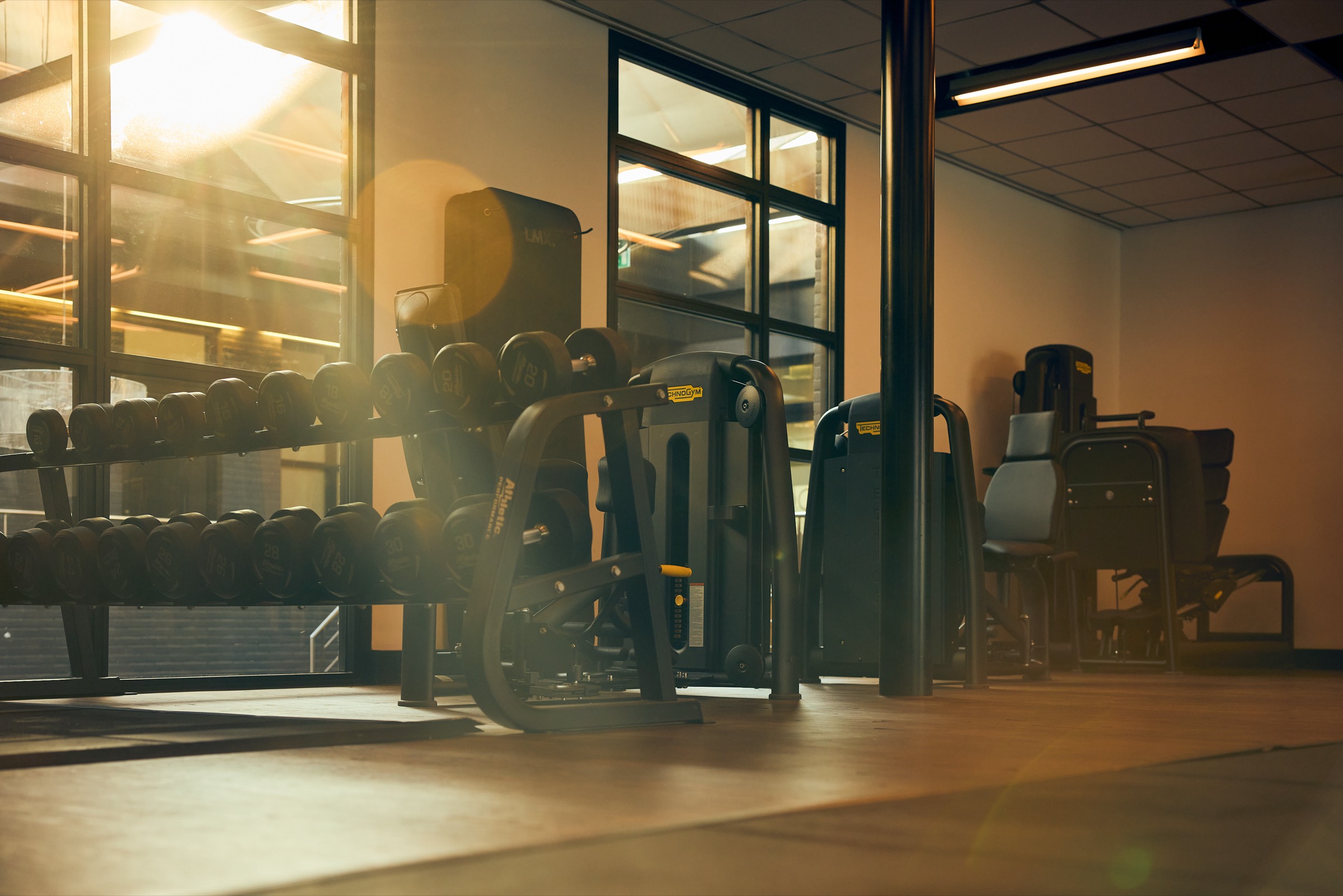 Dumbbellrek en Technogym-toestellen bij de ramen van de trainingsruimte